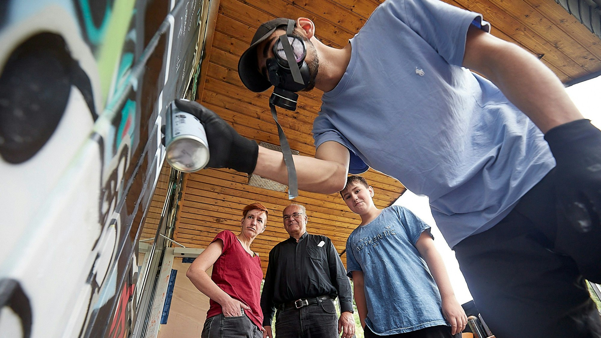 Das Bild zeigt einen Jugendlichen, der, geschützt durch eine Maske, ein Grafitti stpayt. Drei Menschen schauen ihm zu.