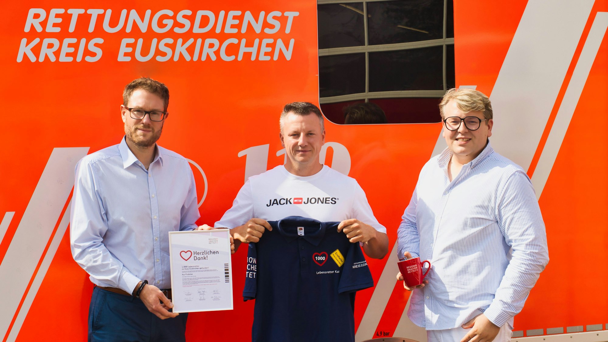 Drei Männer stehen vor einem Rettungswagen. Einer hält eine Urkunde in der Hand, einer ein T-Shirt, einer eine Tasse.