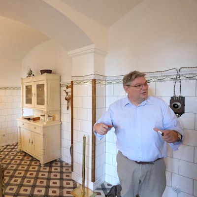 Ein Mann steht in einem Raum mit gefliesten Wänden. In der Küche stehen ein alter Herd und ein alter Küchenschrank,