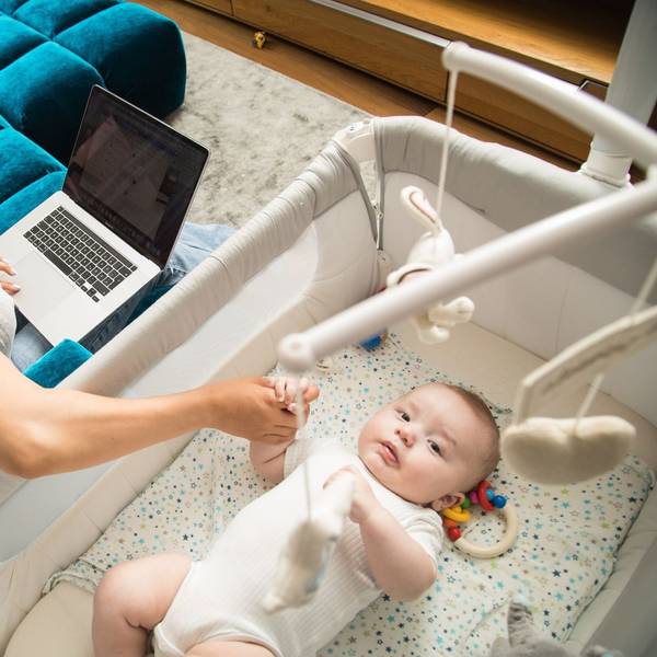 Eine Mutter sitzt auf einer Couch und betreut ihr kleines Baby, auf ihrem Schoß steht ein Laptop.