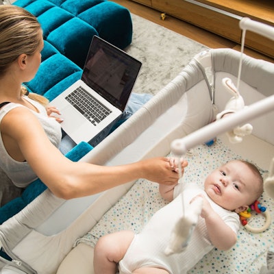 Eine Mutter sitzt auf einer Couch und betreut ihr kleines Baby, auf ihrem Schoß steht ein Laptop.