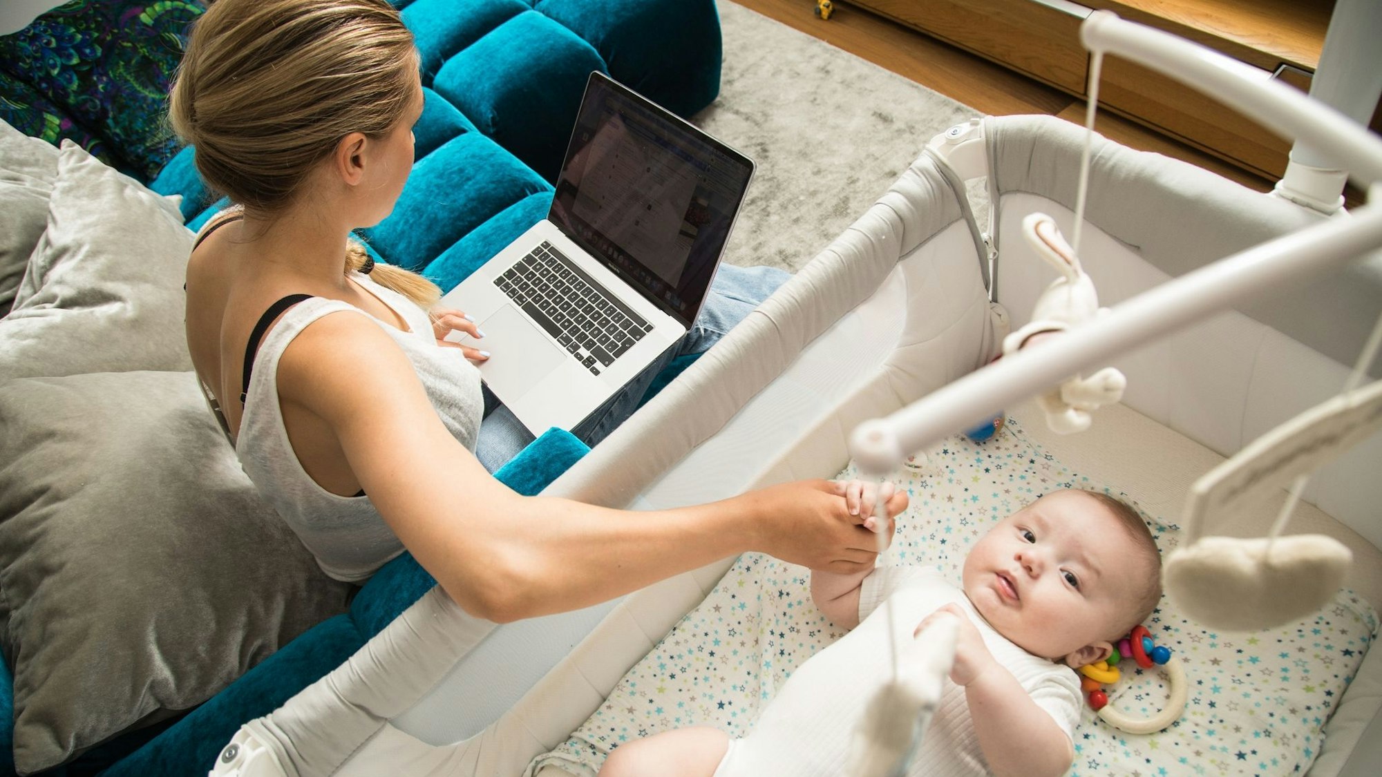 Eine Mutter sitzt auf einer Couch und betreut ihr kleines Baby, auf ihrem Schoß steht ein Laptop.