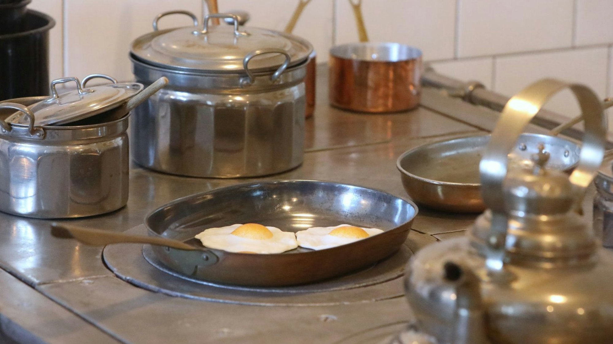 Silberne und kupferne Töpfe, eine alte Bratpfanne mit unechten Spiegeleiern und ein Kessel stehen auf einem alten Herd.