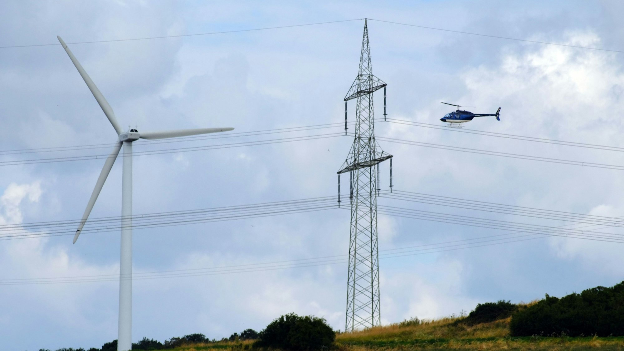 Ein Hubschrauber fliegt nah am Mast einer Stromleitung vorbei. Links neben dem Strommast ist eine Windkraftanlage zu sehen.