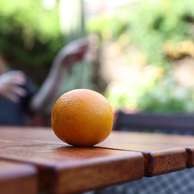 Im Vordergrund liegt eine Orange auf einem Tisch, im Hintergrund ist unscharf ein Mann in schwarzem Shirt zu sehen.