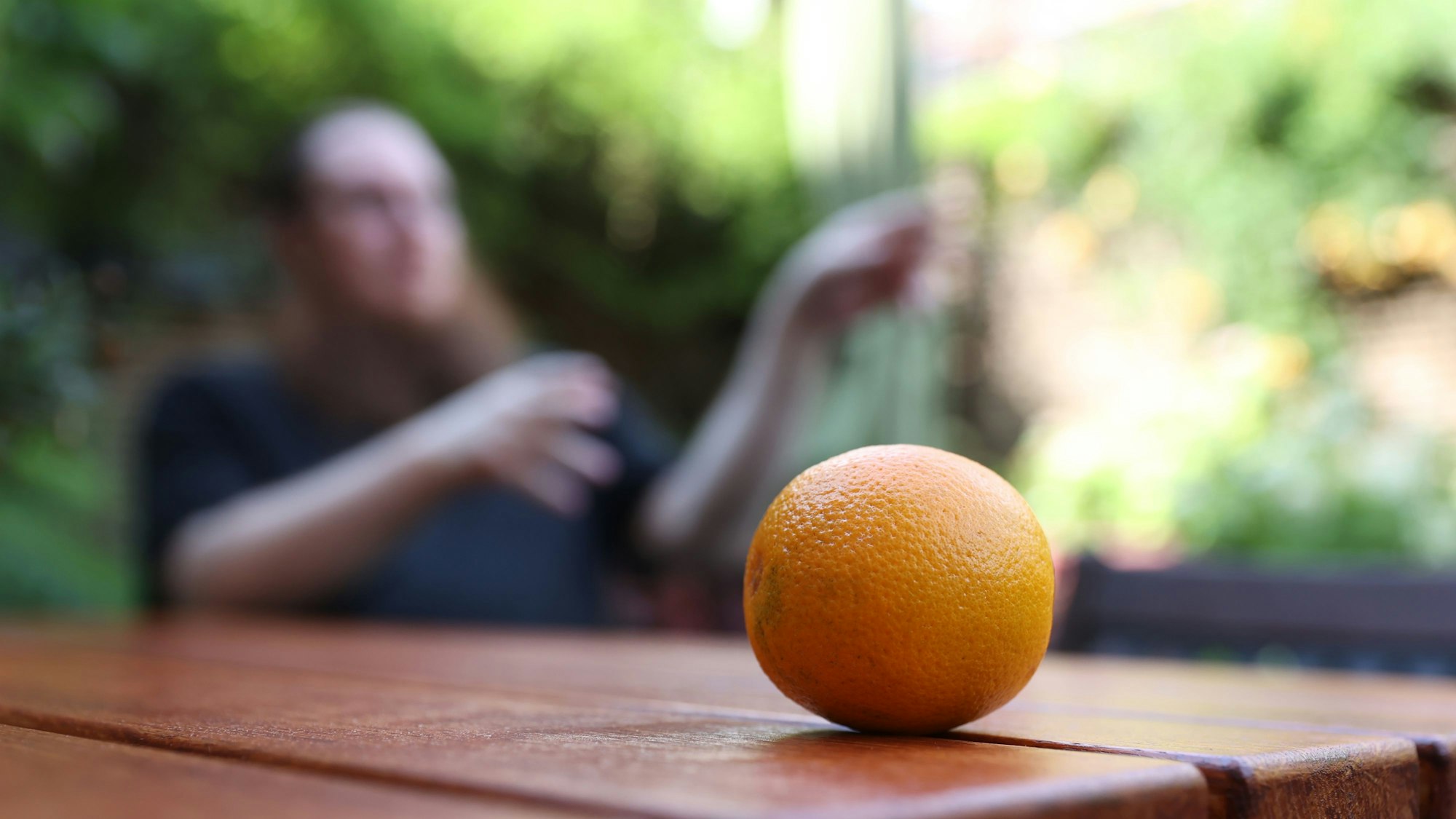 Im Vordergrund liegt eine Orange auf einem Tisch, im Hintergrund ist unscharf ein Mann in schwarzem Shirt zu sehen.