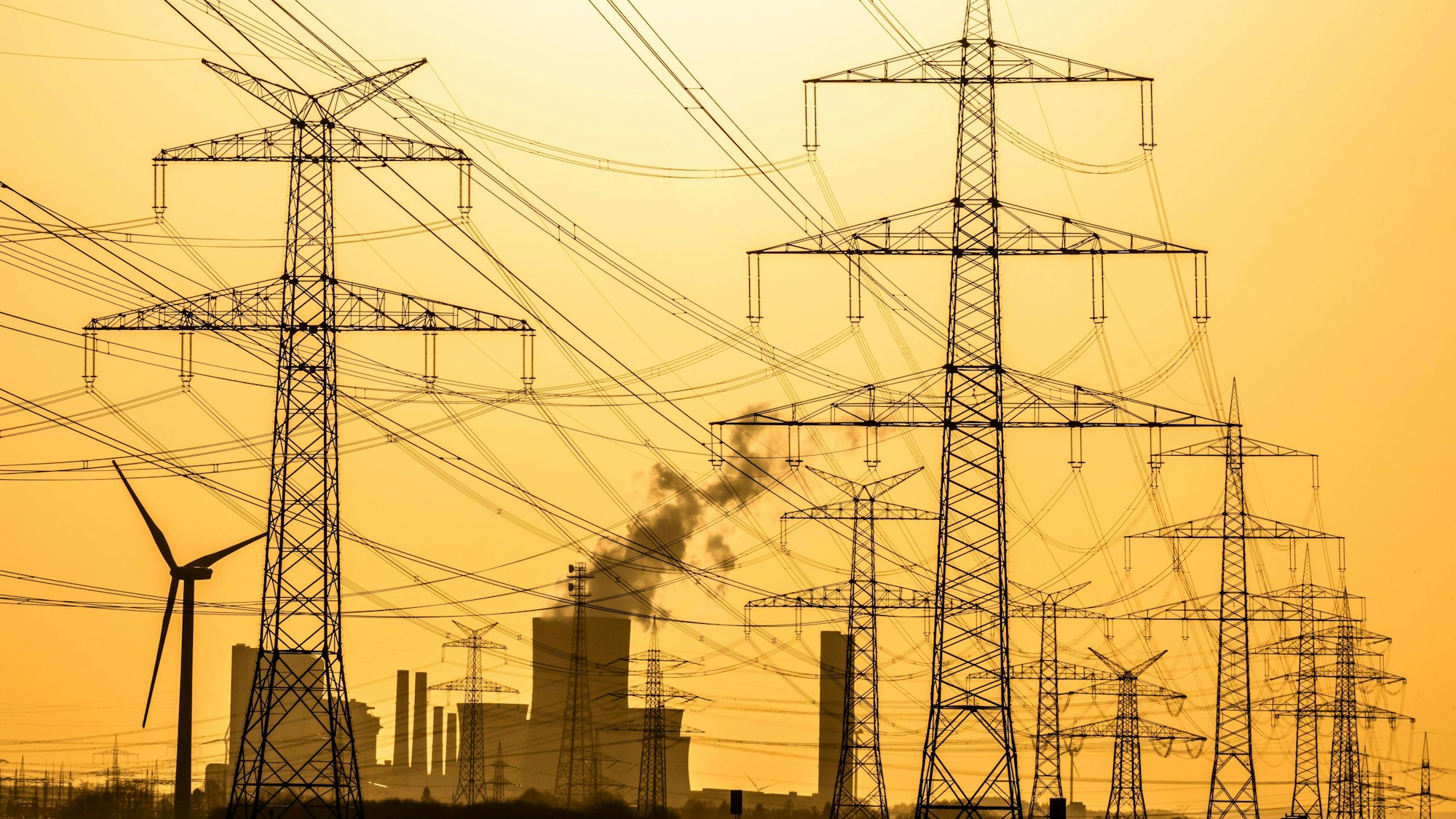 Das Foto zeigt Windräder und Strommasten bei Sonnenuntergang vor dem Braunkohlekraftwerk Niederaußem der RWE Power AG.