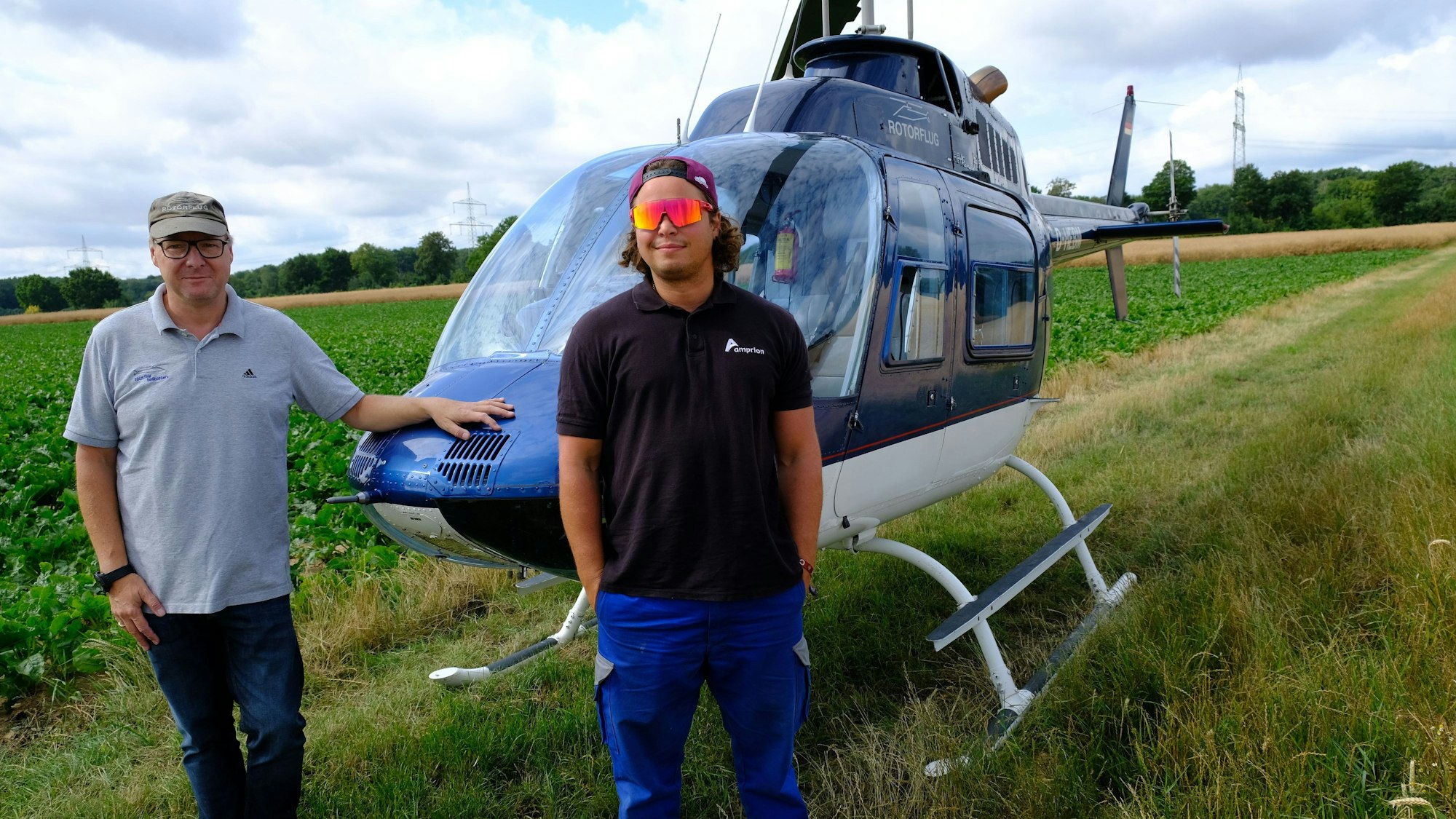 Das Bild zeigt zwei Männer vor einem Hubschrauber, der auf einer Wiese steht.