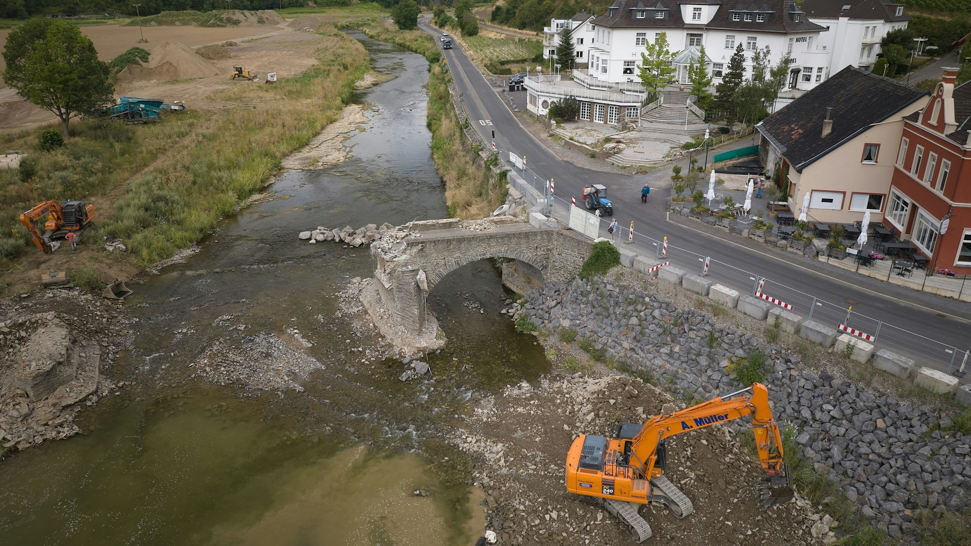Das Bild zeigt einen Blick auf die nur noch mit einem Brückenbogen vorhandene Steinbogenbrücke über dem Fluss Ahr neben einer asphaltierten Straße. Im Vordergrund steht ein orangefarbener Bagger, rechts sind Häuser zu sehen. Das Bild ist aus der Luft, aber aus niedriger Höhe aufgenommen.