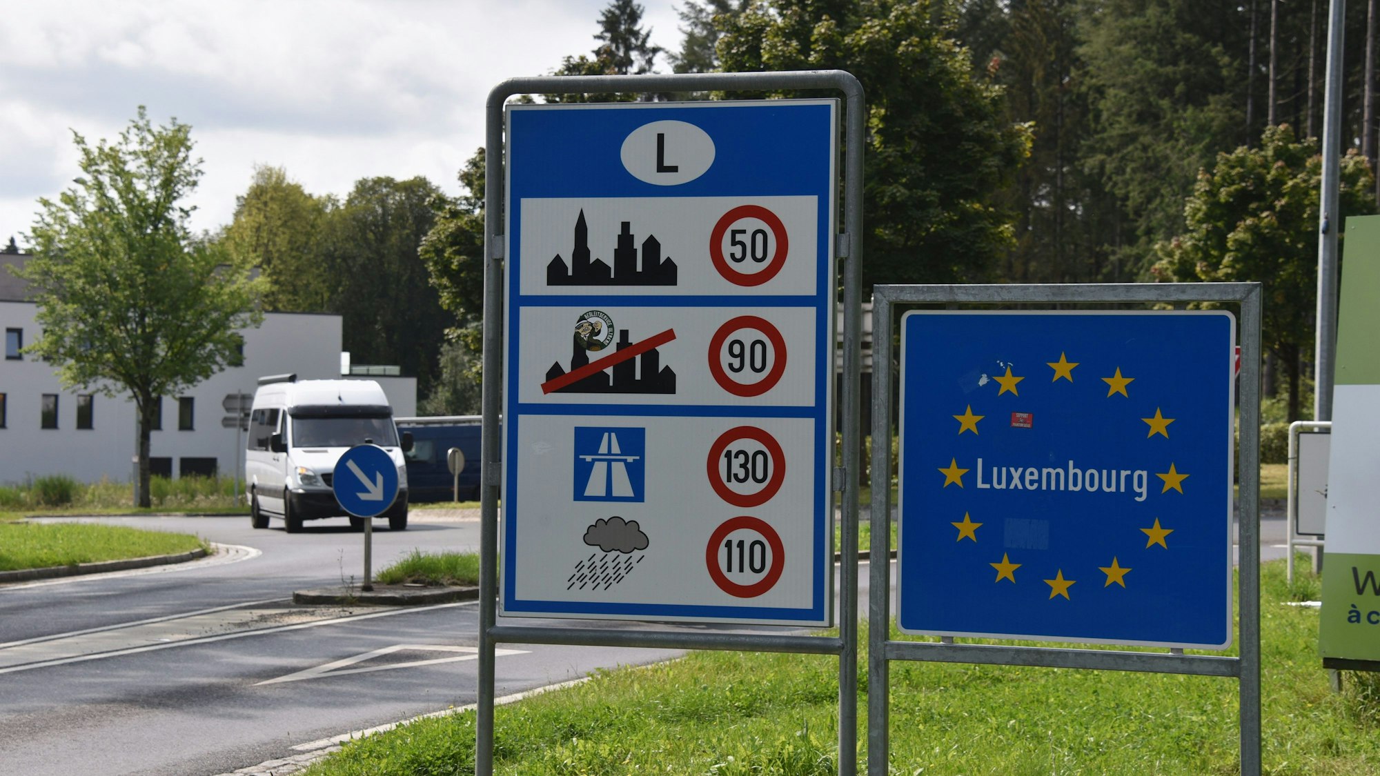 Das Grenzschild von Luxemburg, eingefasst von Europa-Sternen auf blauem Grund, aufgenommen an der deutsch-luxemburgischen Grenze neben einem Schild mit Geschwindigkeitsbegrenzungen.