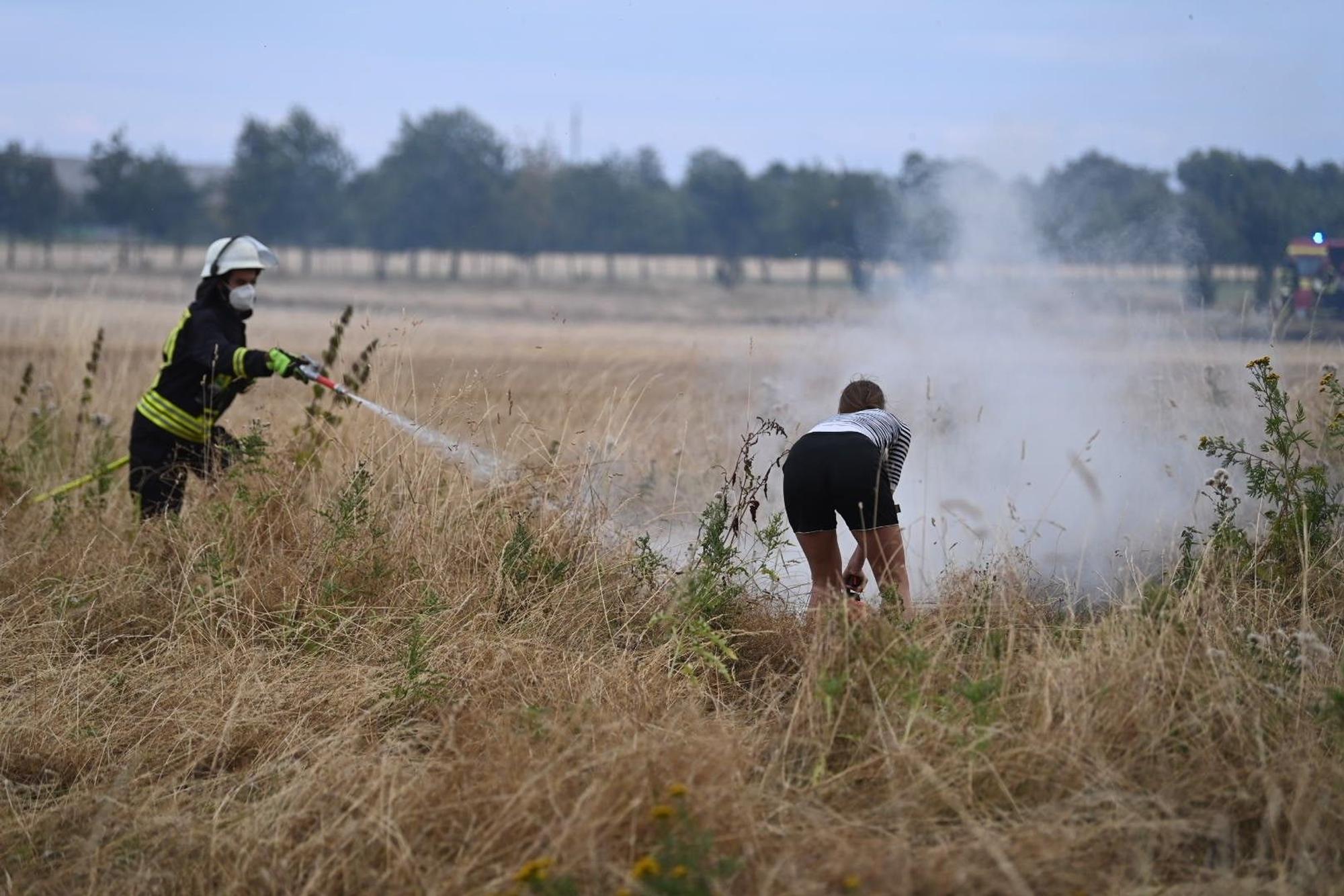 Ein Feuerwehrmann löscht einen Kleinbrand mit dem Strahlrohr, eine Frau unterstützt ihn mit einem Feuerlöscher.