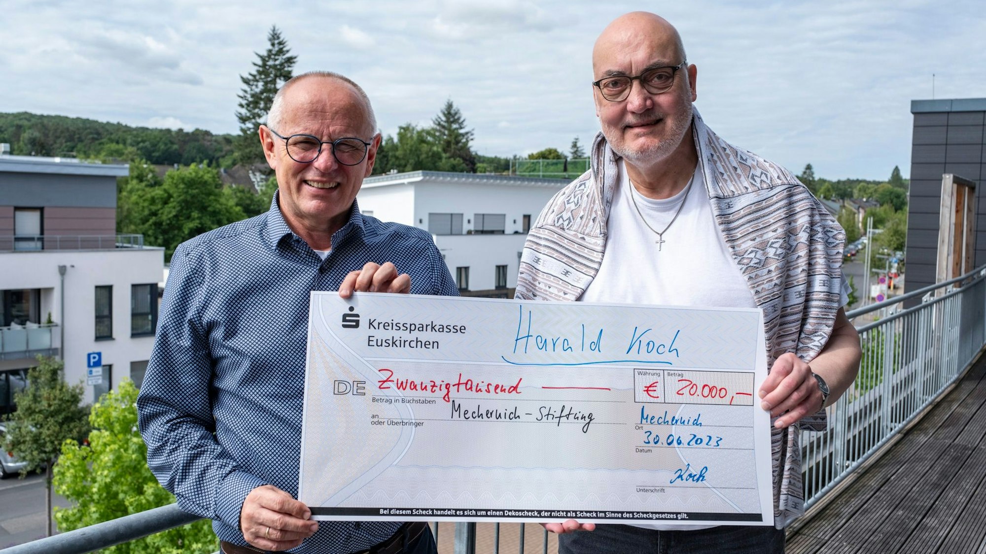 Zwei Männer stehen auf einem Balkon und halten einen Scheck über 20.000 Euro in die Kamera.