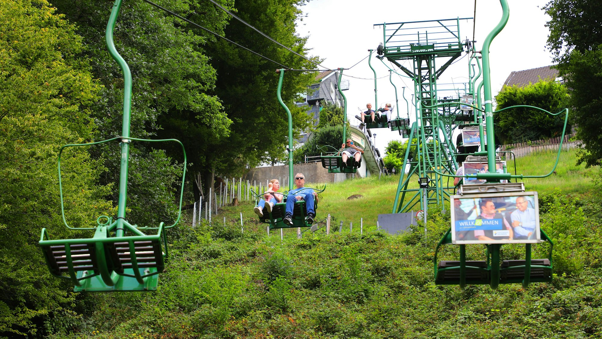 Menschen fahren in einem Sessellift einen begrünten Hang hinauf und hinunter.