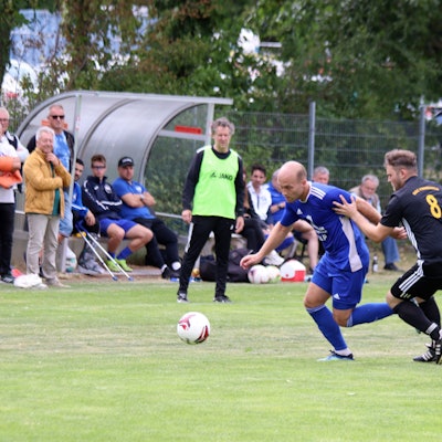 Weilerswists Felix Kortholt zieht an Luca Riedling vorbei. Im Hintergrund beobachten Trainer Horst Bartz im gelben Leibchen und die Zuschauer die Aktion.