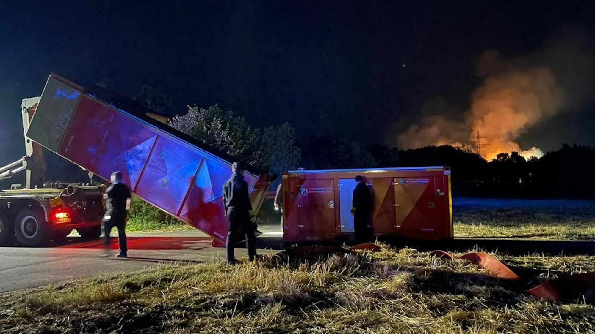 Ein Feuerwehrfahrzeug setzt einen Abrollcontainer ab. Im Hintergrund lodern Flammen.