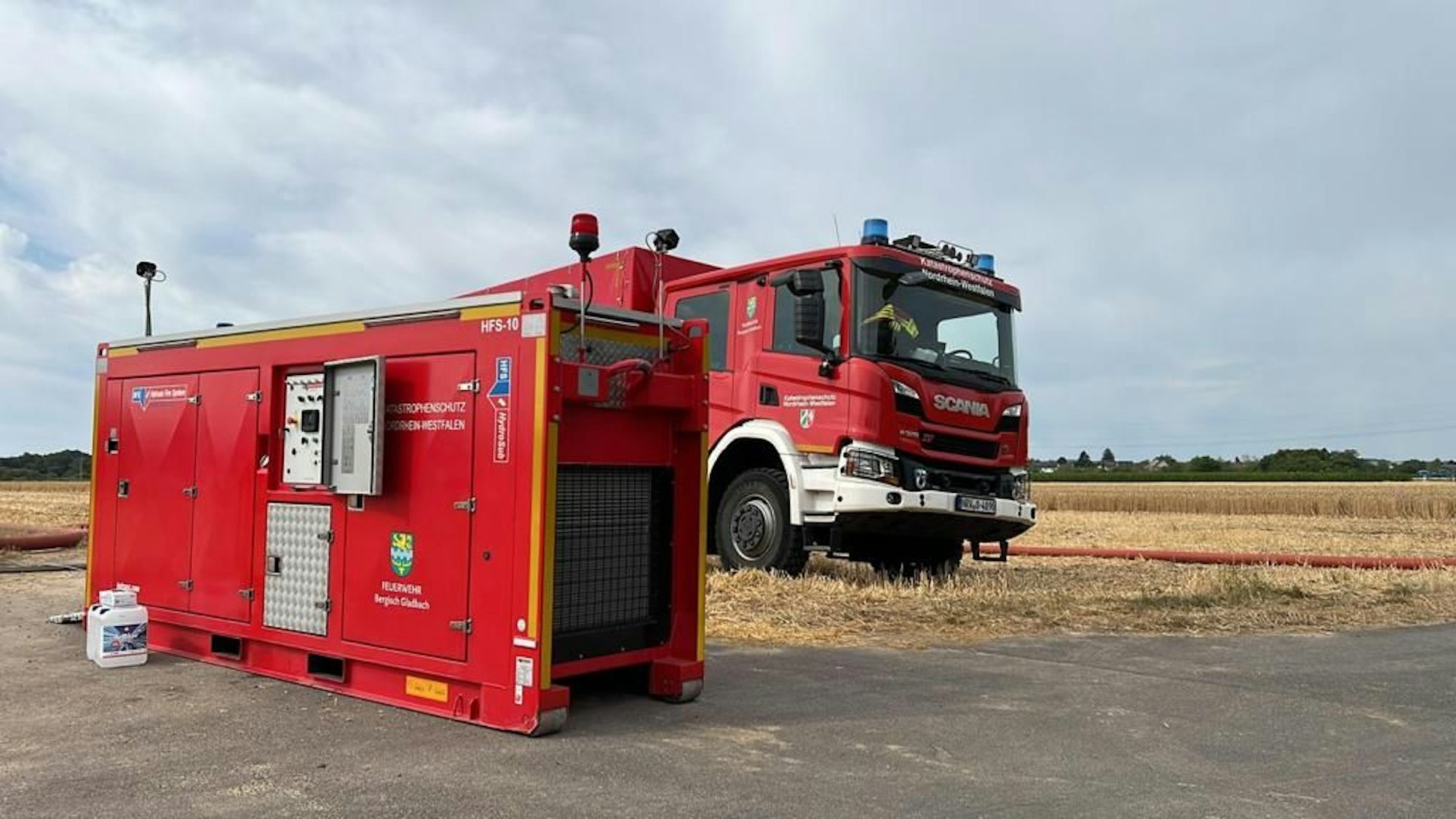 Ein Feuerwehrfahrzeug mit einem Abrollgerät stehen vor einer trockenen Wiesenfläche.