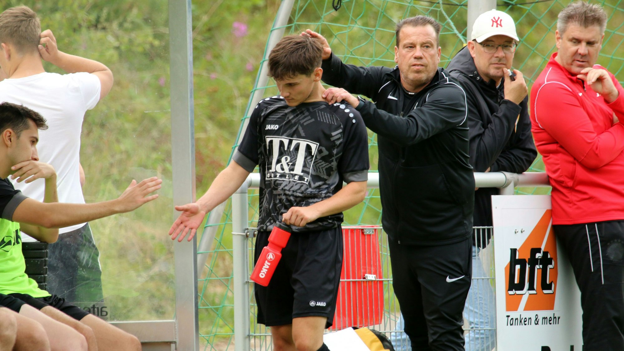 JSG-Trainer Stephan Reimer tröstet den verletzten Nicolas Woywod mit einem Tätscheln des Kopfes. Woywod, in der linken Hand eine rote Trinkflasche, schlägt mit einem Mitspieler ab.