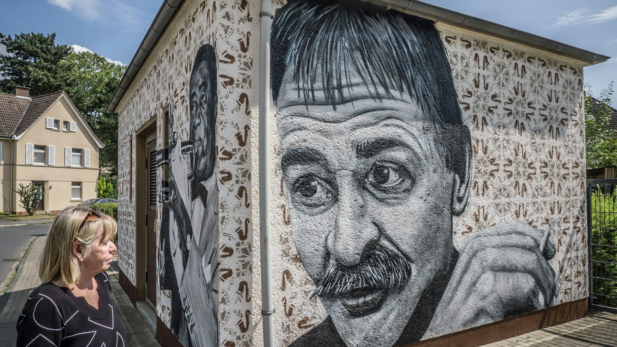 Ingrid Orth guckt sich das Porträt ihres verstorbenen Ehemanns, Wolfgang Orth, auf einem Strom-Häuschen nach einem Bild von Ralf Krieger aus dem Leverkusener Anzeiger. Auf der Frontseite: Louis Armstrong nach diesem Bild: https://www.loc.gov/pictures/item/00652636/ Der Fassadenmaler: Street Art-Künstler Jens Lagemann alias ArtByOpus