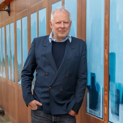 Der Schauspieler Axel Milberg lehnt an einer Hauswand, er trägt Jeans, einen dunkelblauen Pullover und ein blaues Sakko.