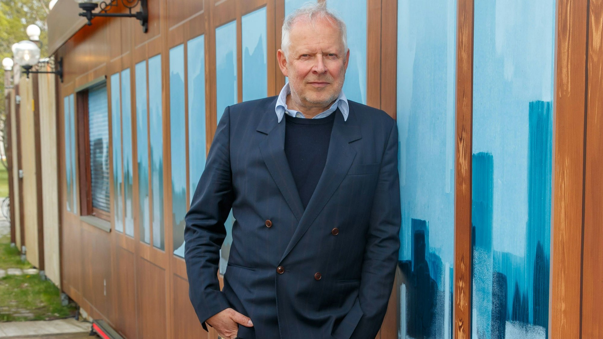 Der Schauspieler Axel Milberg lehnt an einer Hauswand, er trägt Jeans, einen dunkelblauen Pullover und ein blaues Sakko.
