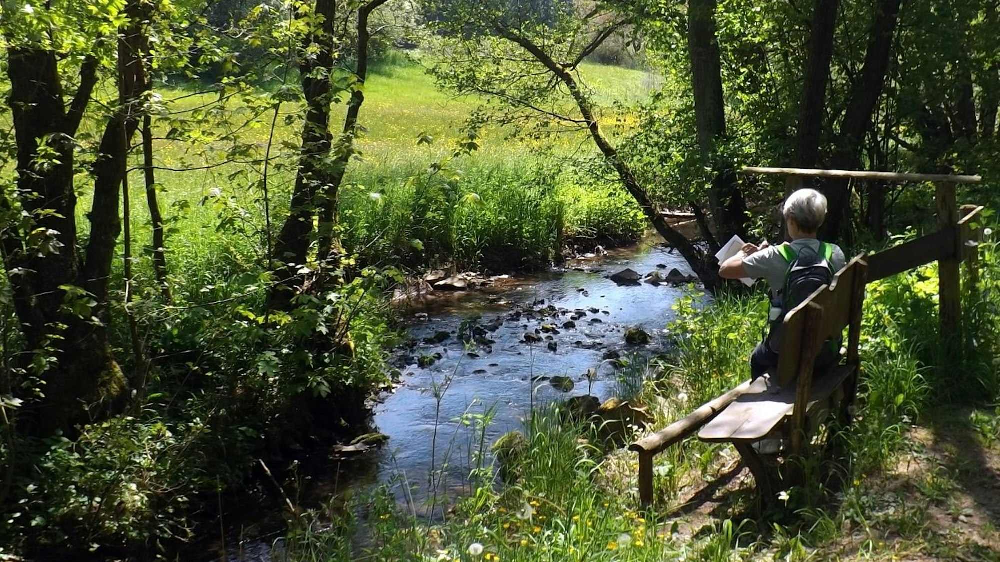 Eine Frau sitzt auf einer Bank und schaut in eine Wanderkarte, vor ihr fließt ein idyllischer Bach