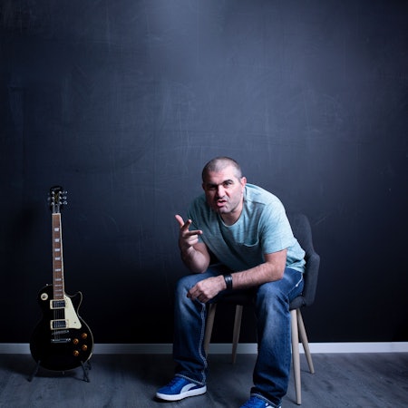 Ein Mann sitzt in einem leeren Raum auf einem Stuhl. Neben ihm steht eine E-Gitarre auf dem Boden.