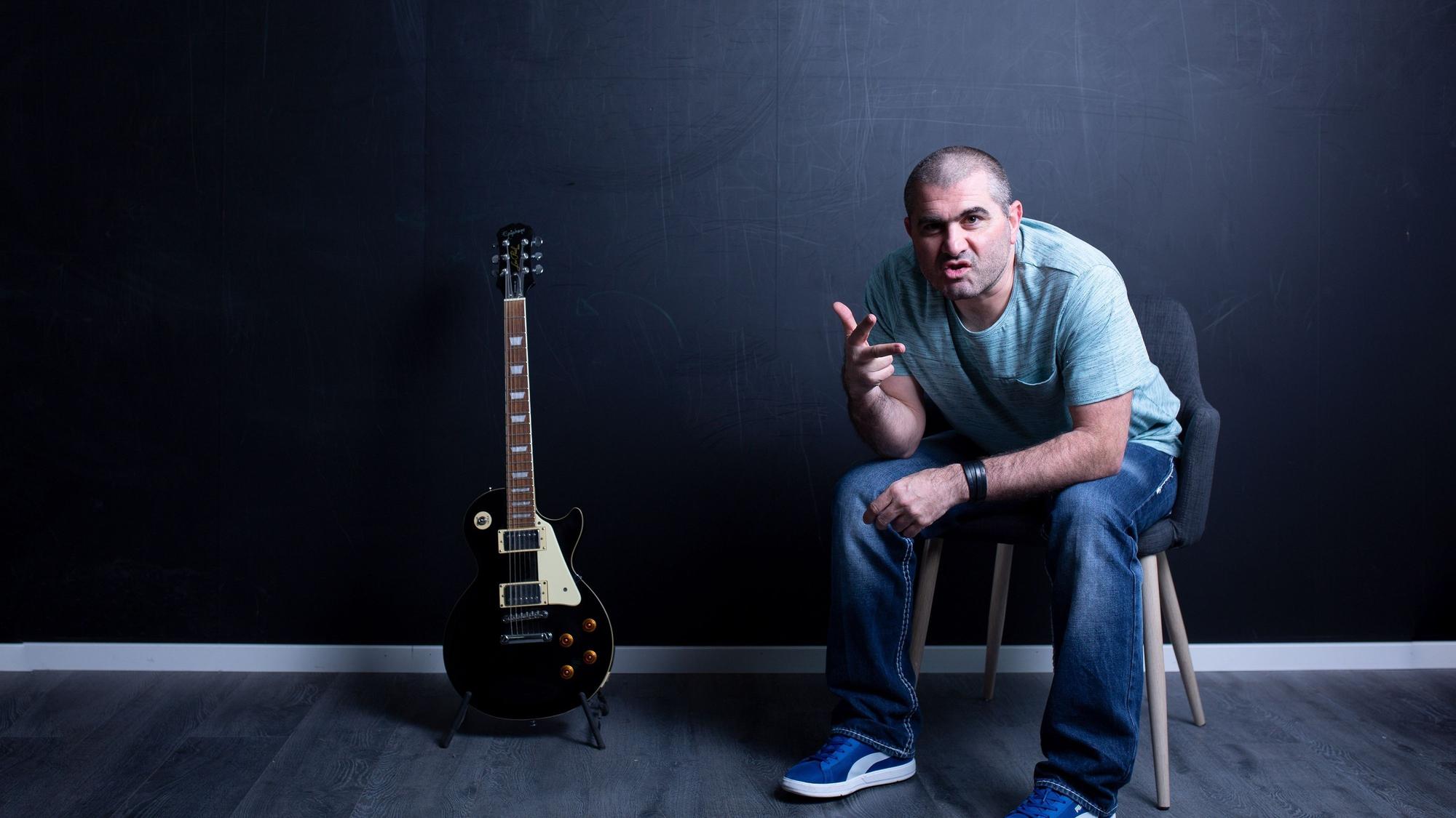 Ein Mann sitzt in einem leeren Raum auf einem Stuhl. Neben ihm steht eine E-Gitarre auf dem Boden.