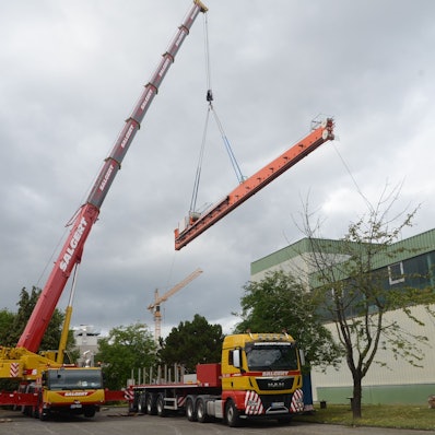 Ein großer Autokran hebt ein langes orange lackiertes Metallteile über einen Tieflader und eine Fabrikhalle hinweg.