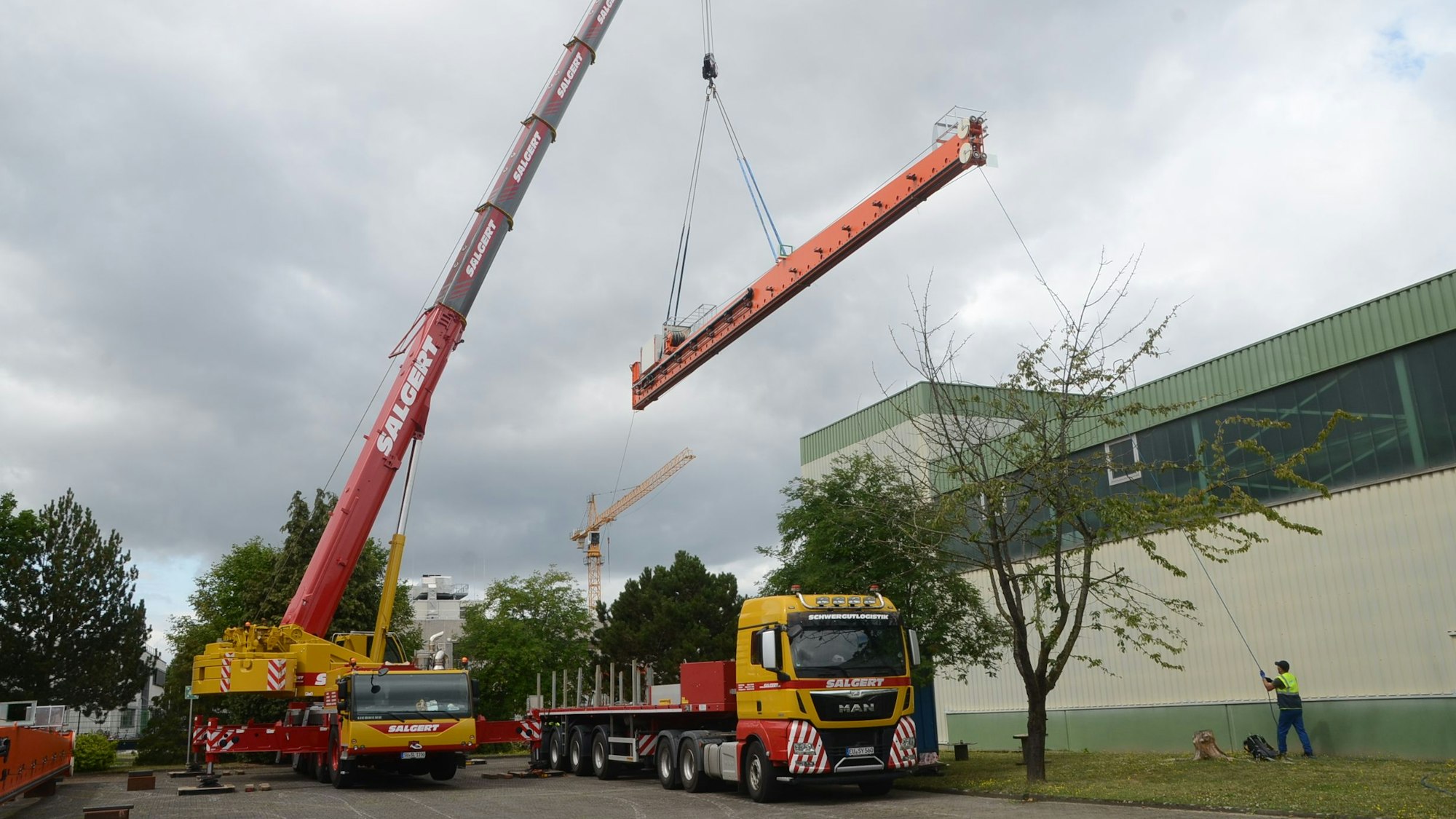Ein großer Autokran hebt ein langes orange lackiertes Metallteile über einen Tieflader und eine Fabrikhalle hinweg.