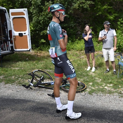 Der Kölner Radprofi Nils Politt vom Team Bora-Hansgrohe steht auf der 19. Etappe der Tour de France nach einem Defekt an seinem Fahrrad am Straßenrand. Im Hintergrund filmen ihn zwei Zuschauer am Straßenrand.
