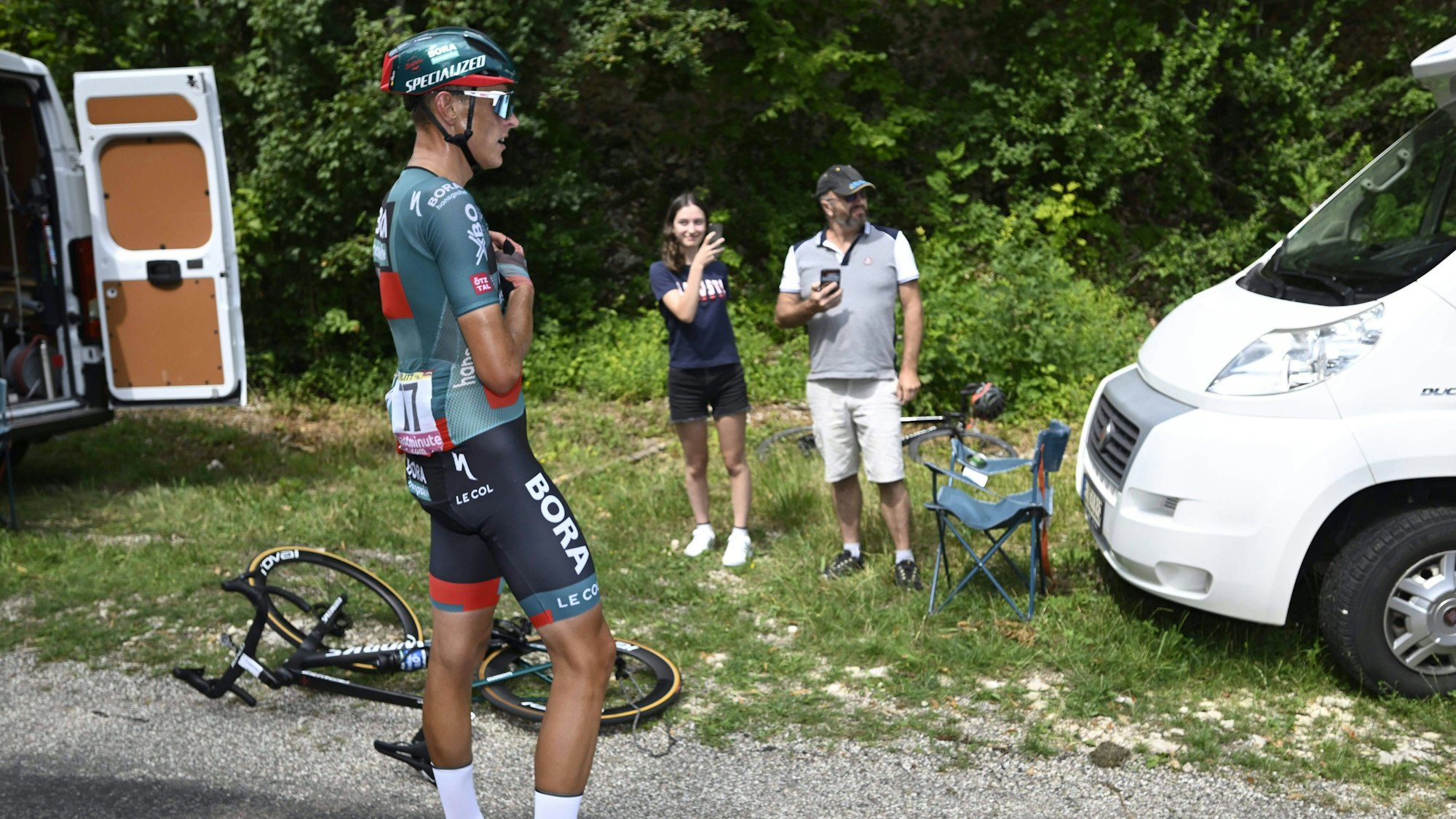 Der Kölner Radprofi Nils Politt vom Team Bora-Hansgrohe steht auf der 19. Etappe der Tour de France nach einem Defekt an seinem Fahrrad am Straßenrand. Im Hintergrund filmen ihn zwei Zuschauer am Straßenrand.