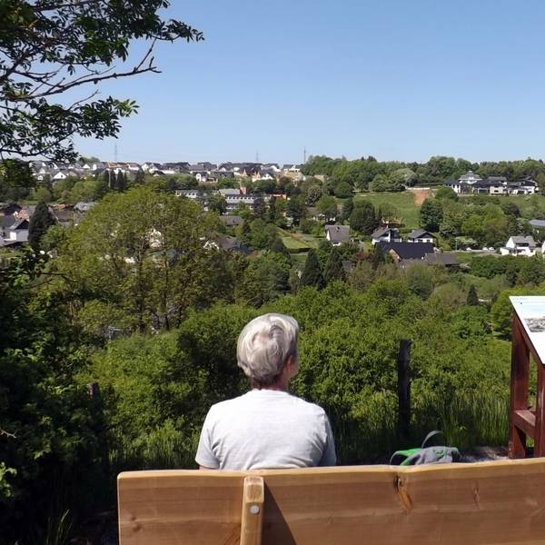Eine Frau sitzt auf einer Bank mit Blick auf ein Dorf im Grünen und pausiert bei einer Wanderung