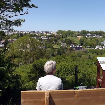 Eine Frau sitzt auf einer Bank mit Blick auf ein Dorf im Grünen und pausiert bei einer Wanderung