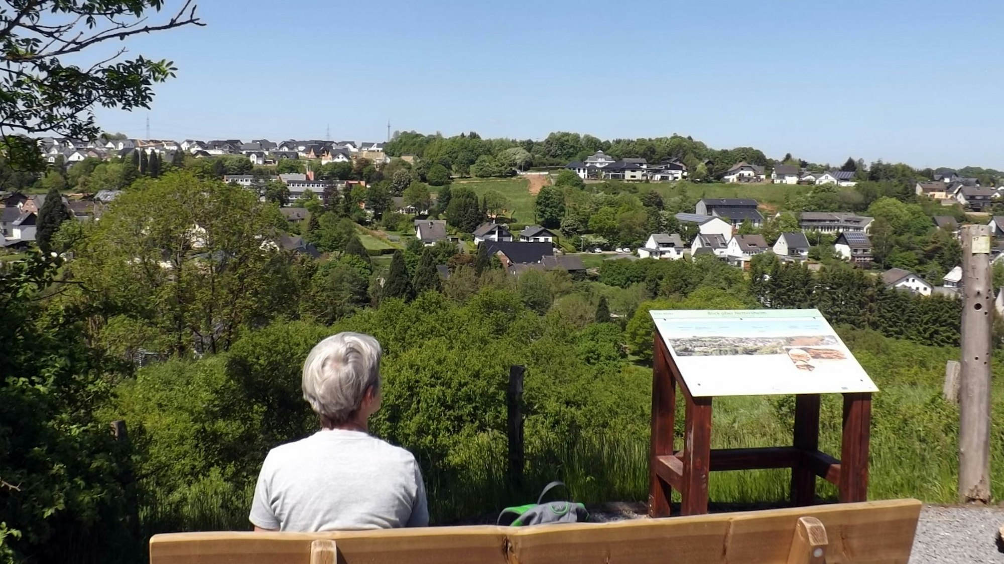 Eine Frau sitzt auf einer Bank mit Blick auf ein Dorf im Grünen und pausiert bei einer Wanderung