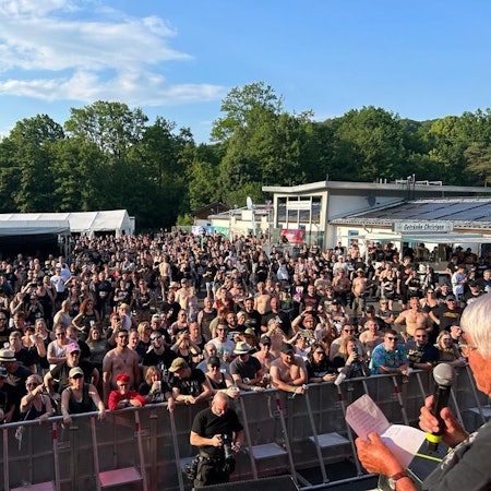 Eine Frau spricht auf der Bühne zum Publikum. Sie hält ein Mikrofon und ein Blatt Papier in der Hand.