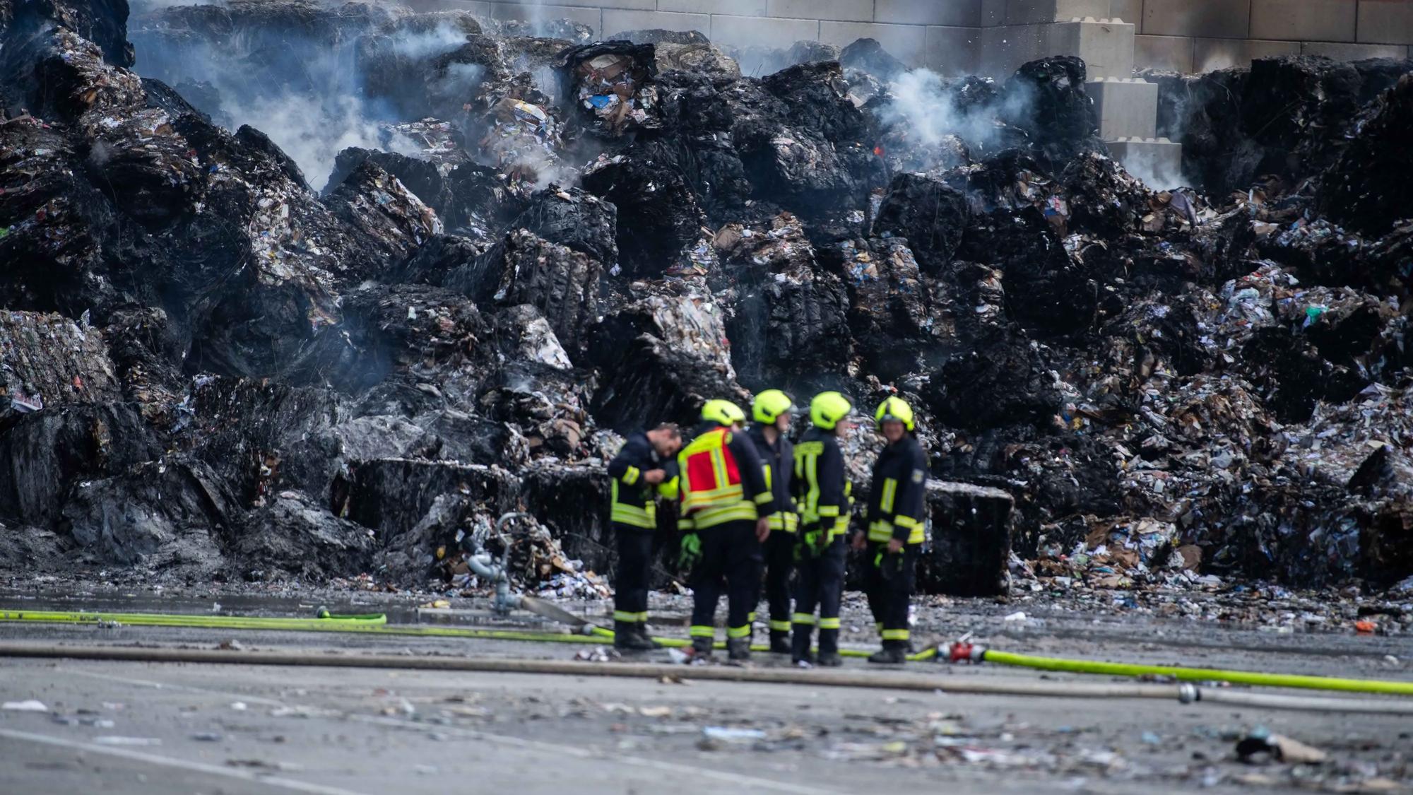 Mehrere Feuerwehrleute stehen vor einer großen Menge abgebrannter Papierballen.