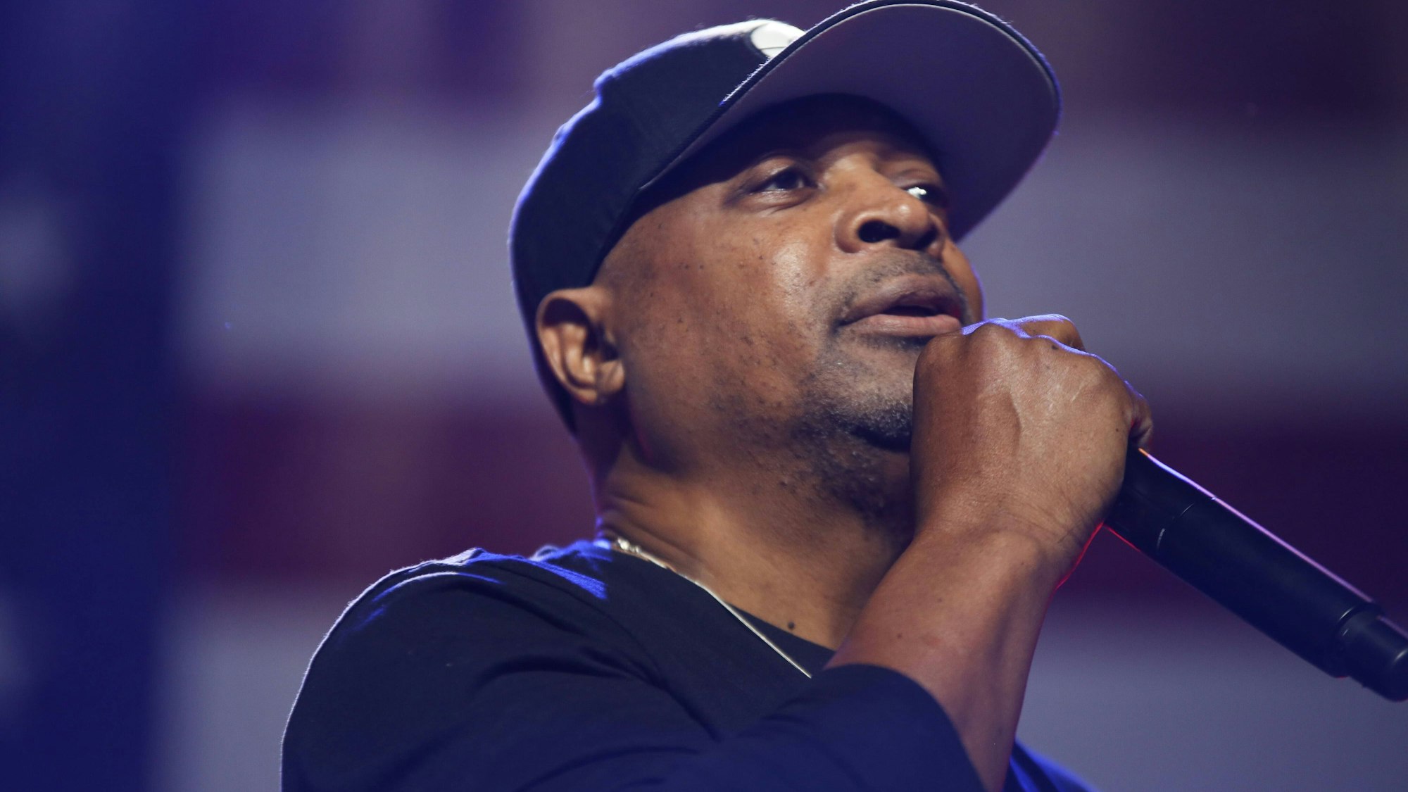 Chuck D singt in ein Mikrofon. Er hat eine dunkle Mütze und einen dunklen Pullover an, der Hintergrund ist verschwommen und violett.
