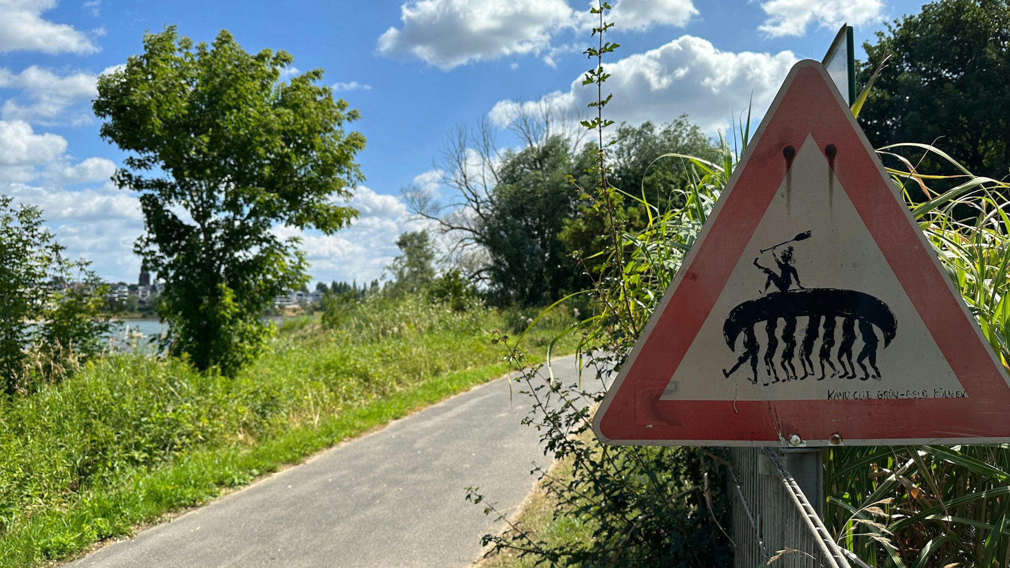An einem Rhein-Uferweg im Kölner Süden steht ein Verkehrsschild, auf dem sieben Menschen ein Kanu tragen.