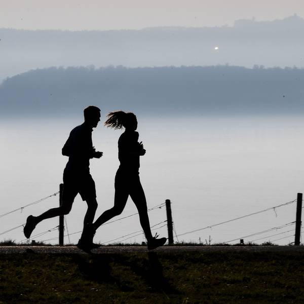 Jogger laufen auf einem Wanderweg in Richtung Meersburg, während im Hintergrund die Stadt Konstanz am Bodensee im Dunst zu sehen ist.
