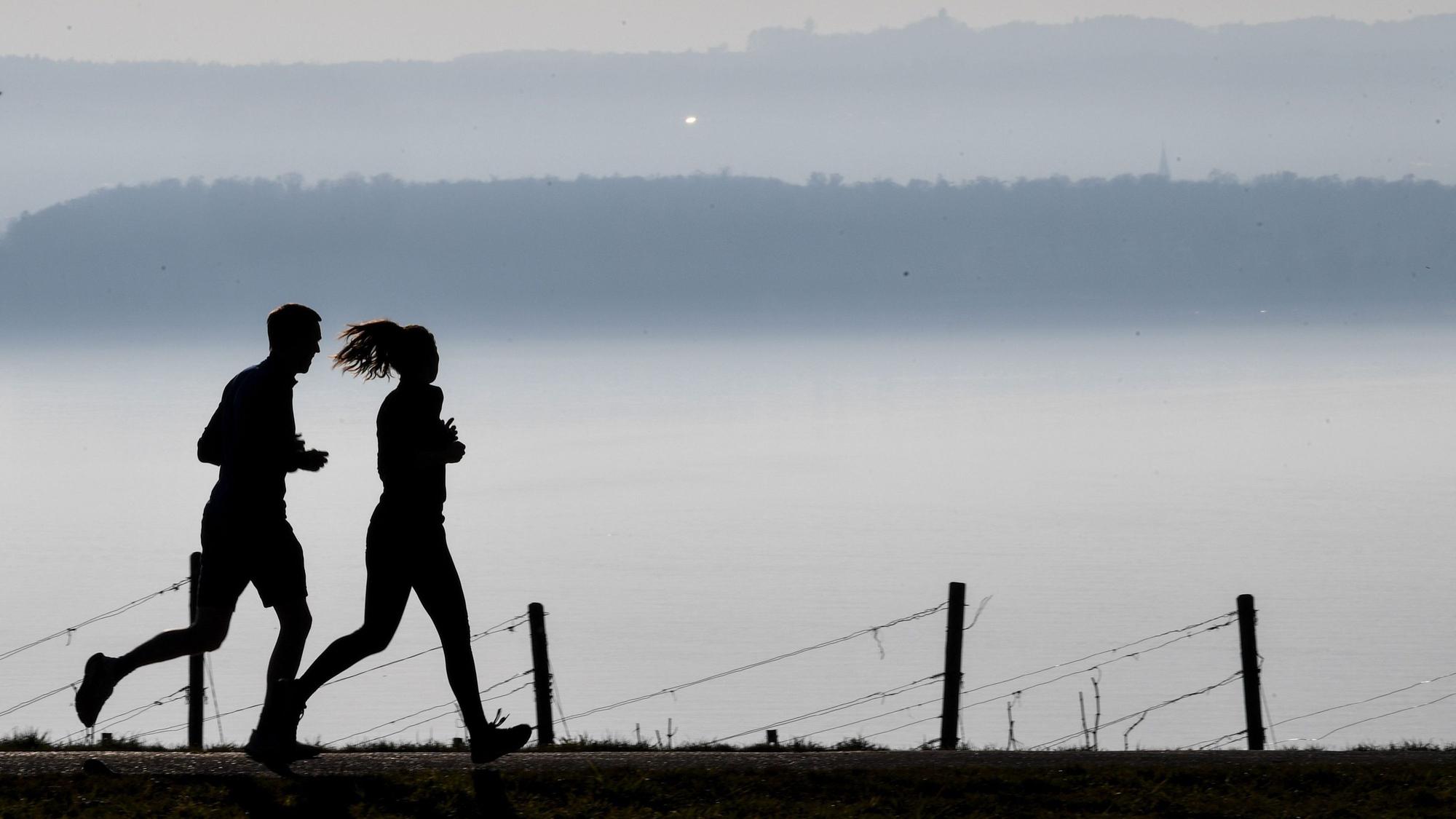 Jogger laufen auf einem Wanderweg in Richtung Meersburg, während im Hintergrund die Stadt Konstanz am Bodensee im Dunst zu sehen ist.