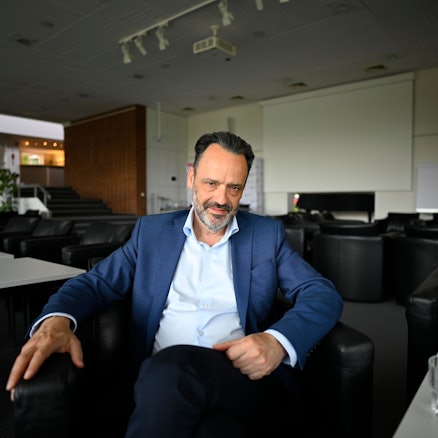 Ein Mann im Anzug sitzt in einem Konferenzraum auf einem Stuhl und schaut in die Kamera.