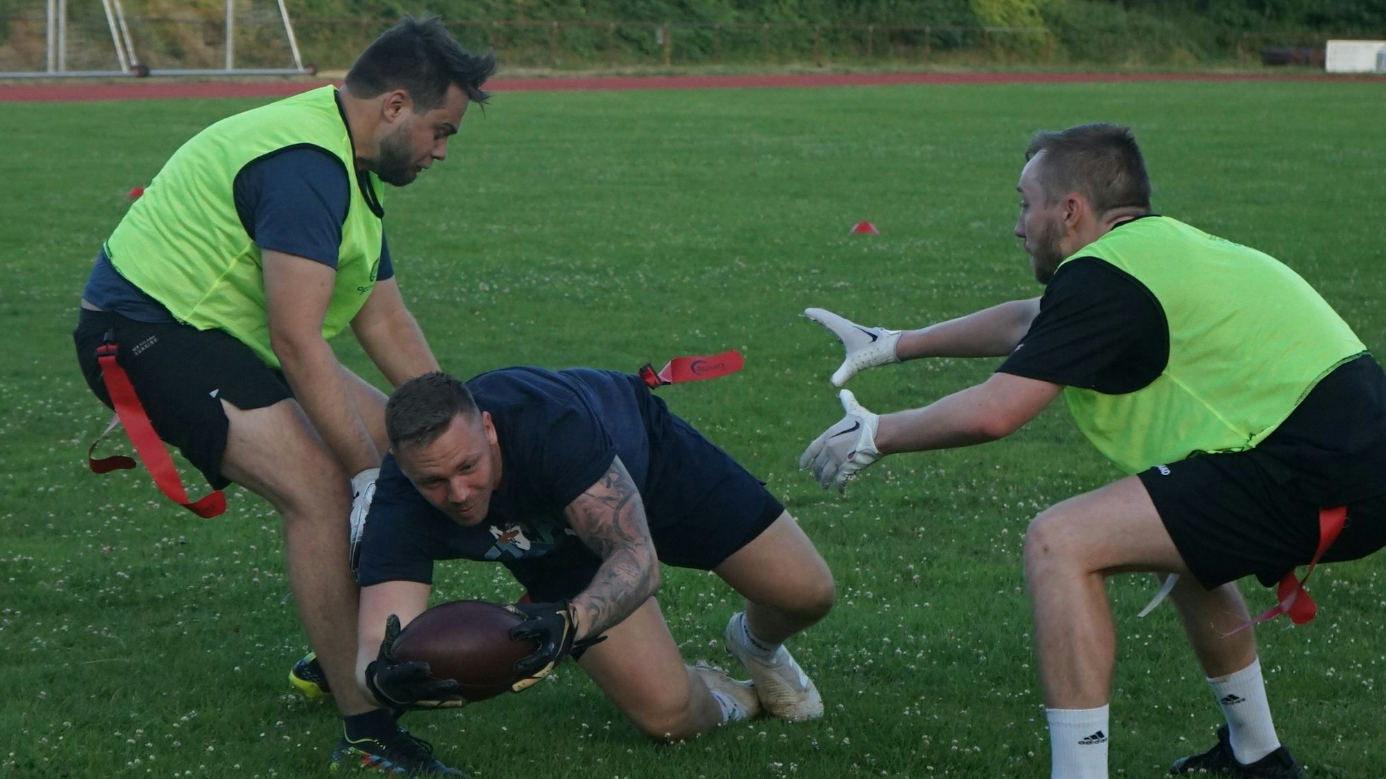 Raccoons-Teammitglied Marvin Bongartz wirft sich mit einem Football auf den Boden. Zwei andere Spieler mit gelben Westen versuchen, ihm die Flaggen aus dem Gürtel zu ziehen.