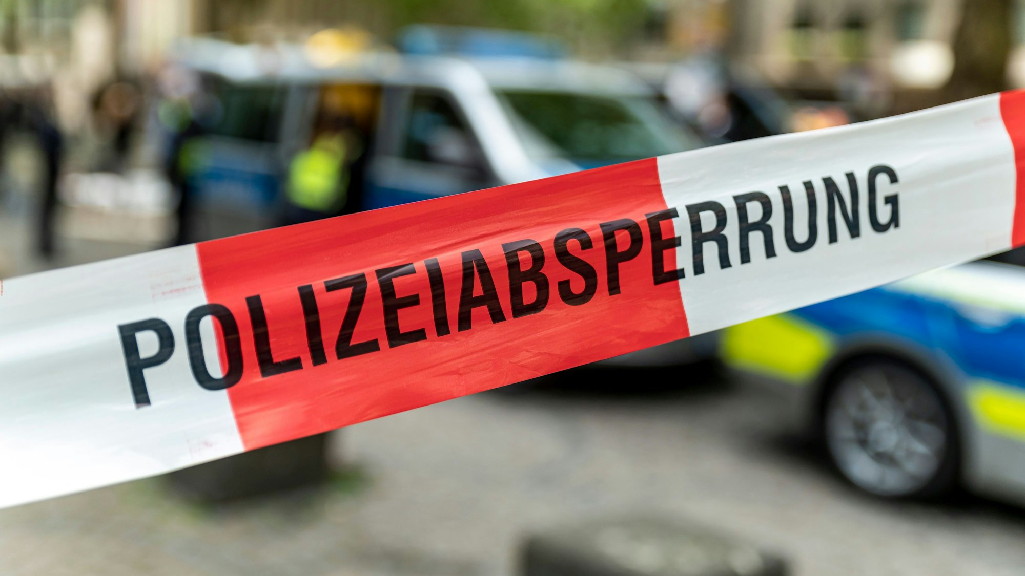 Symbolfoto: Die Aufschrift „Polizeiabsperrung“ steht auf einem Flatterband in den Farben weiß und rot. Dahinter ist unscharf ein Streifenwagen der Polizei zu sehen.