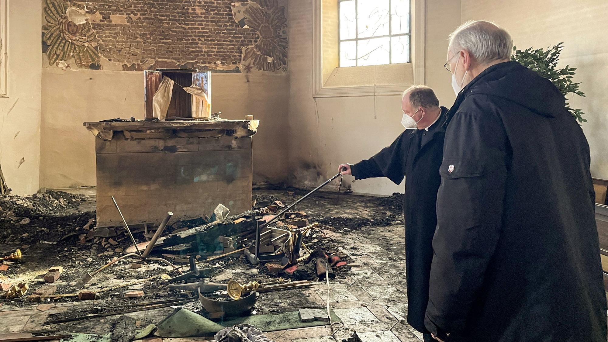 Zwei Priester stehen in einem Kircheninnenraum vor Überrresten nach einem Brand.