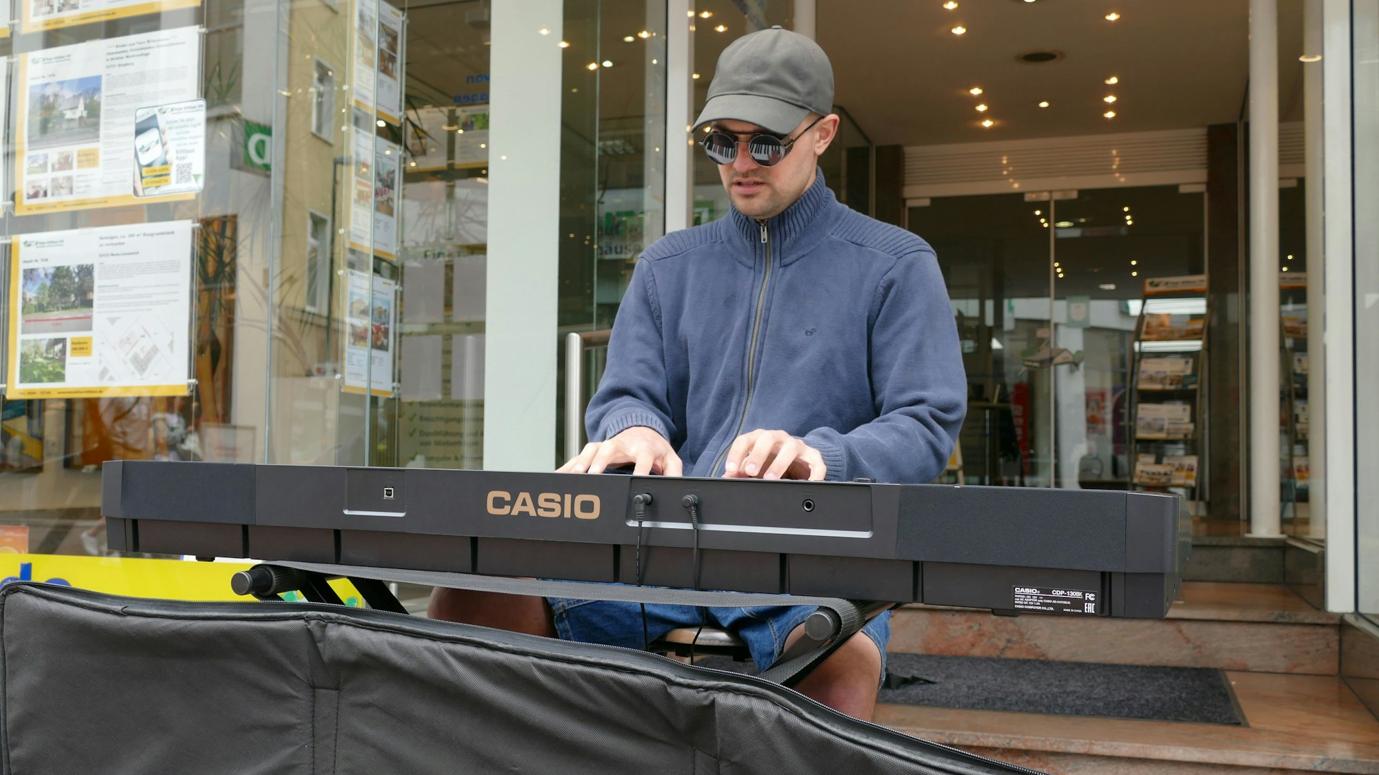 Ein junger Mann mit Baseballkappe, Sonnenbrille und einer blauen Fleecejacke spielt Keyboard vor dem Eingang zu einem Geschäft.