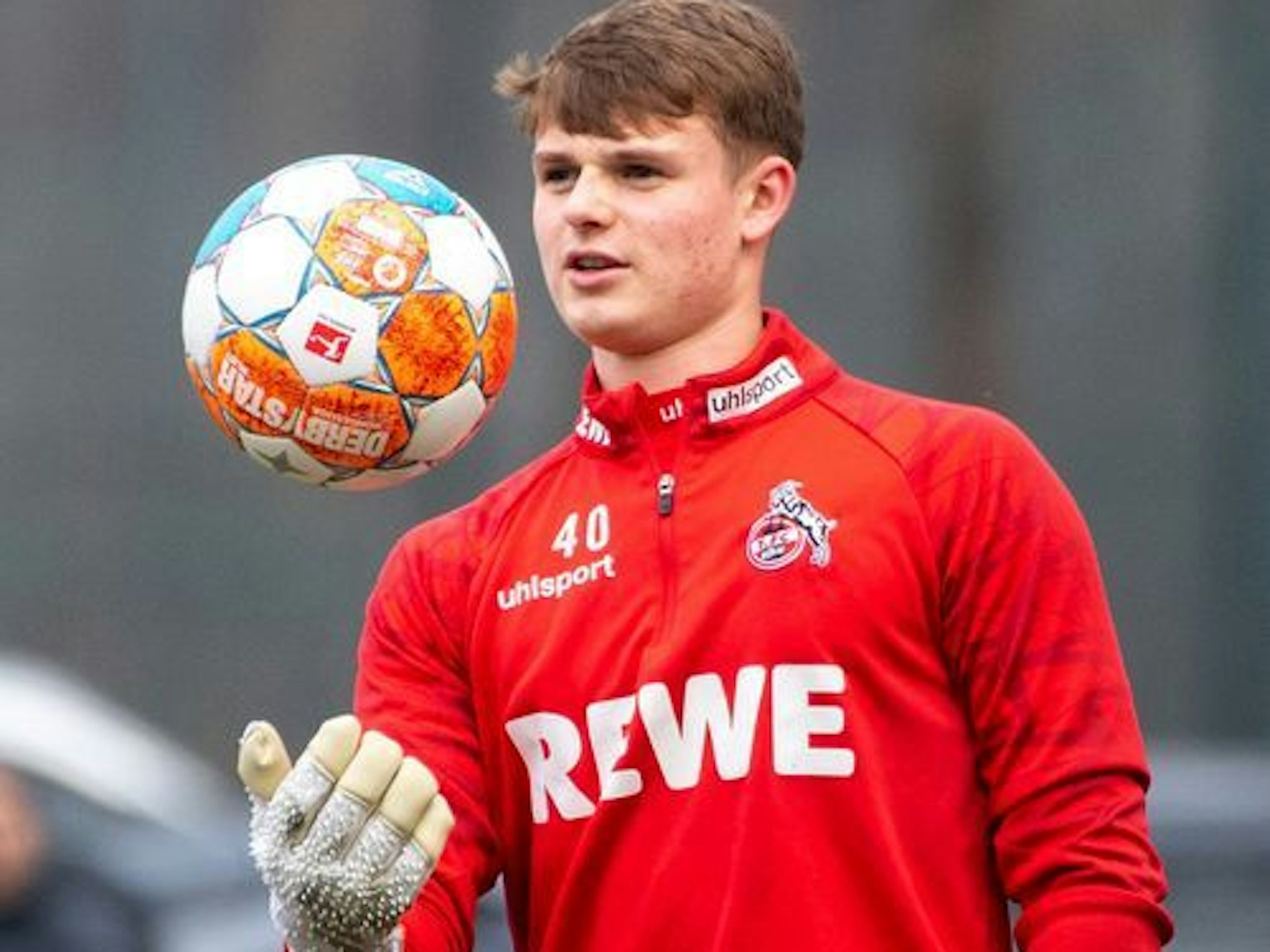 Jonas Urbig steht in einem Trainingsshirt des 1. FC Köln auf dem Fußballplatz und wirft einen Ball hoch.