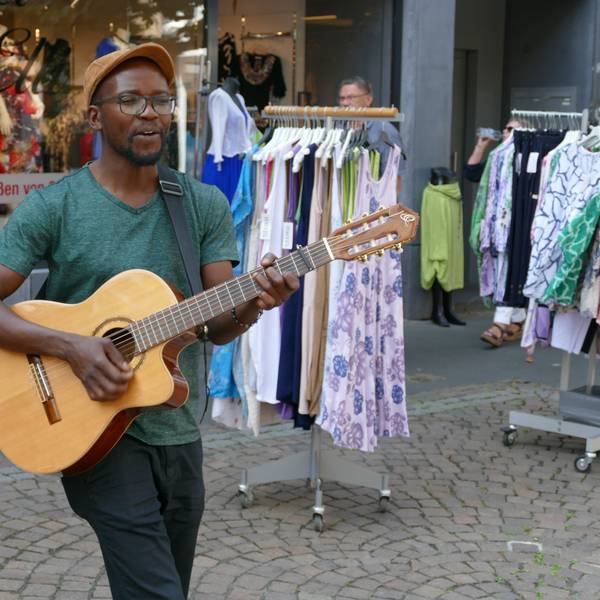 Ein junger Mann mit Schirmmütze und Gitarre; er trägt eine Brille und hat eine dunkle Hautfarbe.
