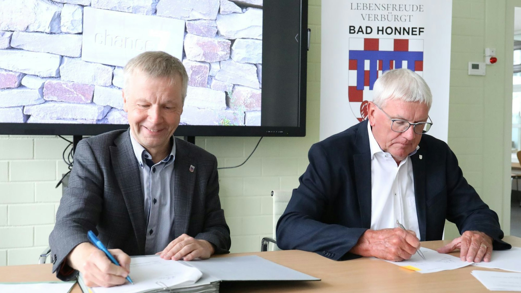 Zwei Männer sitzen nebeneinander an einem Tisch und unterschreiben die Verträge. Im Hintergrund das Stadtlogo mit dem Motto „Lebensfreude verbürgt“.