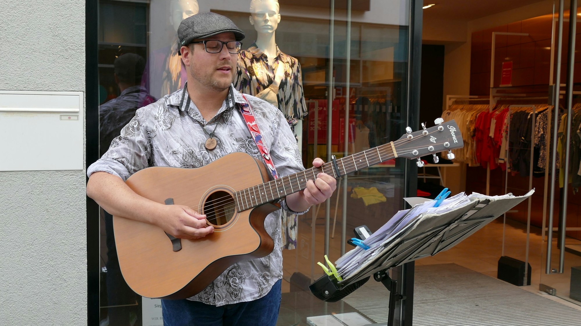 Ein Mann mit Brille und Schirmmütze spielt Gitarre vor dem Eingang eines Geschäfts. Auf seinem Notenständer hat er die Noten mit Wäscheklammern fixiert.