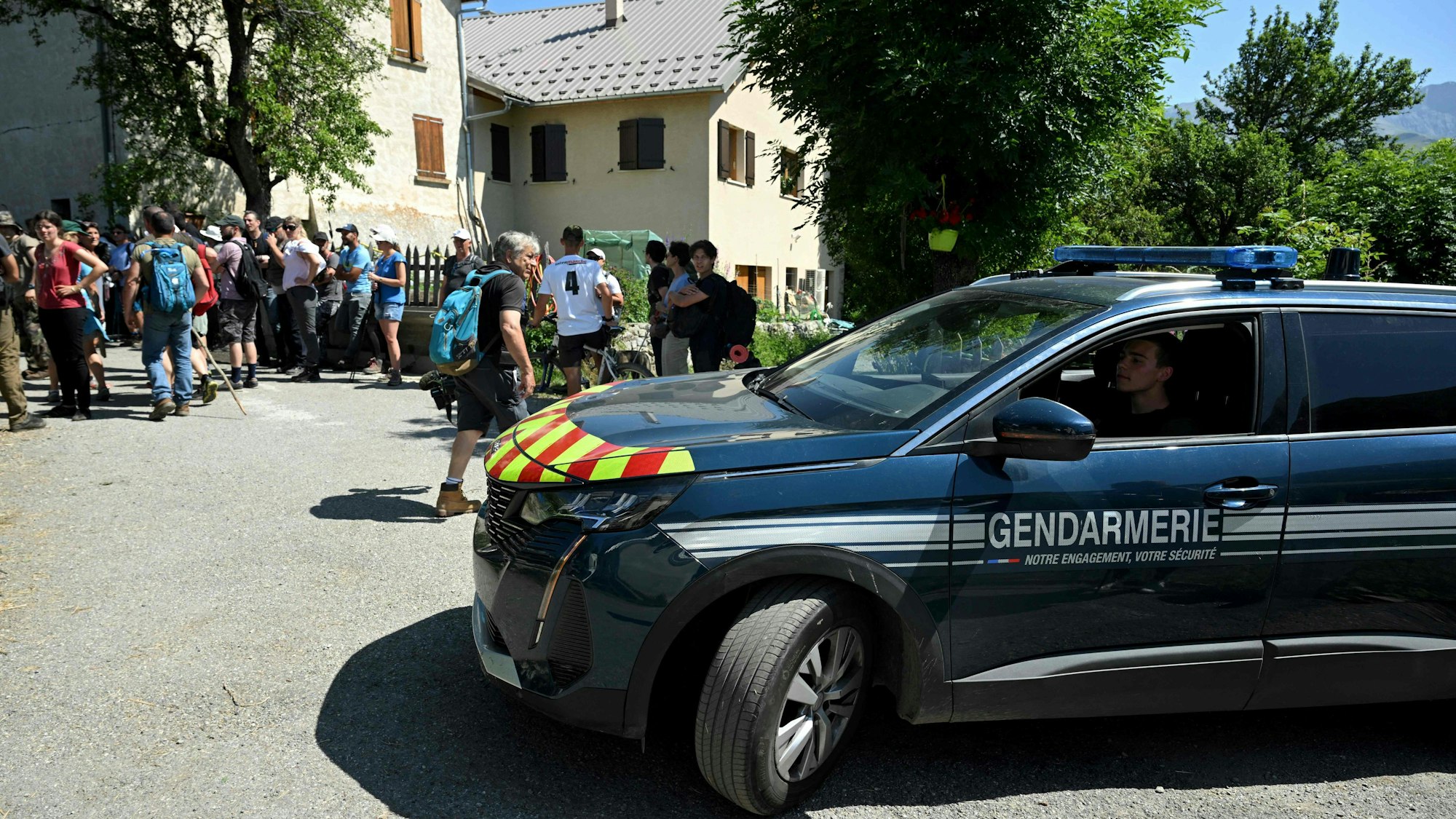 Ein französischer Polizeiwagen der Gendarmerie fährt durch den französischen Ort Le Vernet. Im Hintergrund stehen zahlreiche Helfer, die nach dem vermissten Émile suchen.
